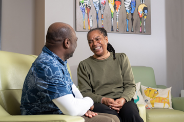 A man and a woman sitting on a living room sofa, smiling and having a conversation.